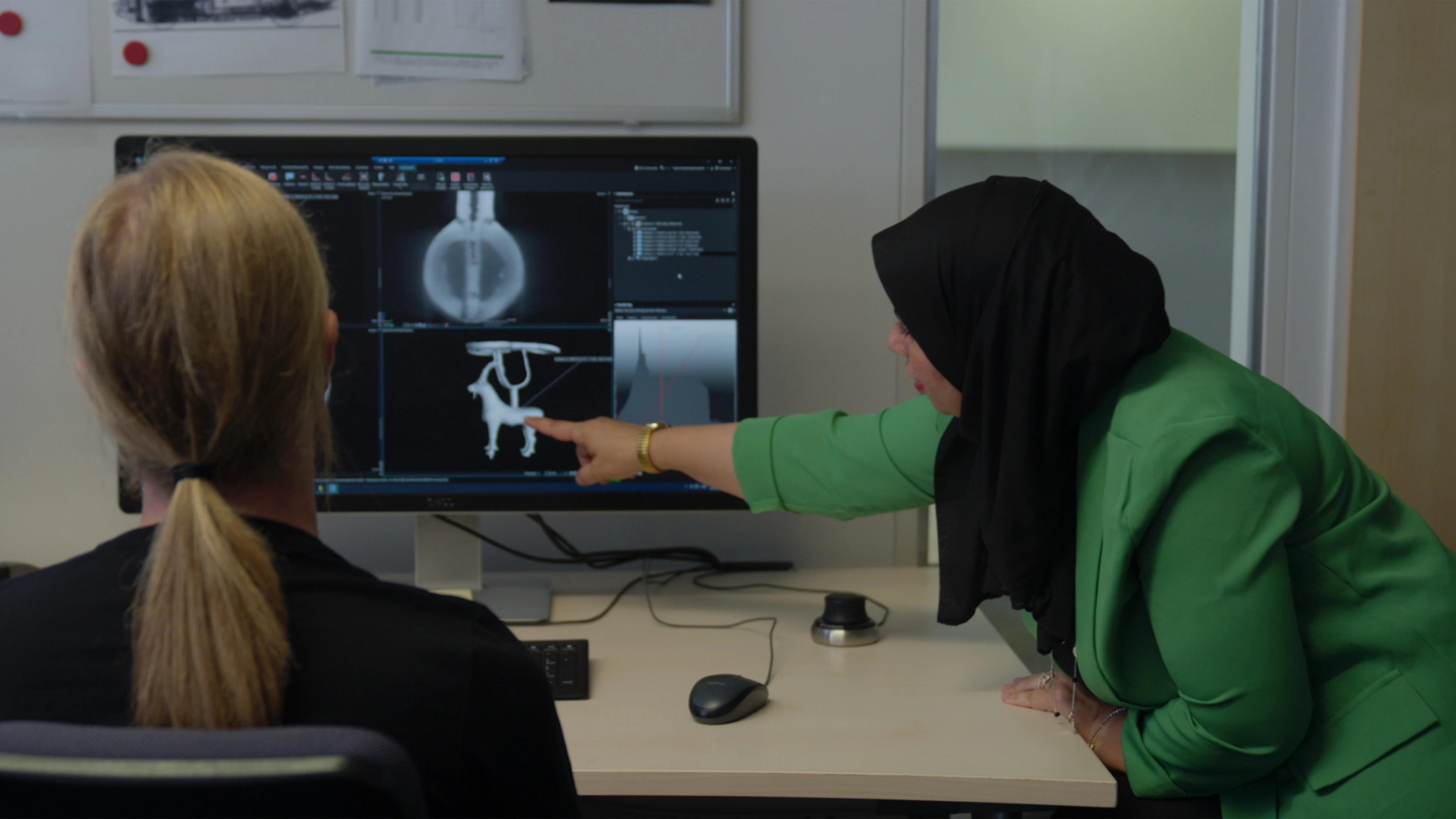 Two people at a desk with a large computer monitor. One person points to an image on the screen.