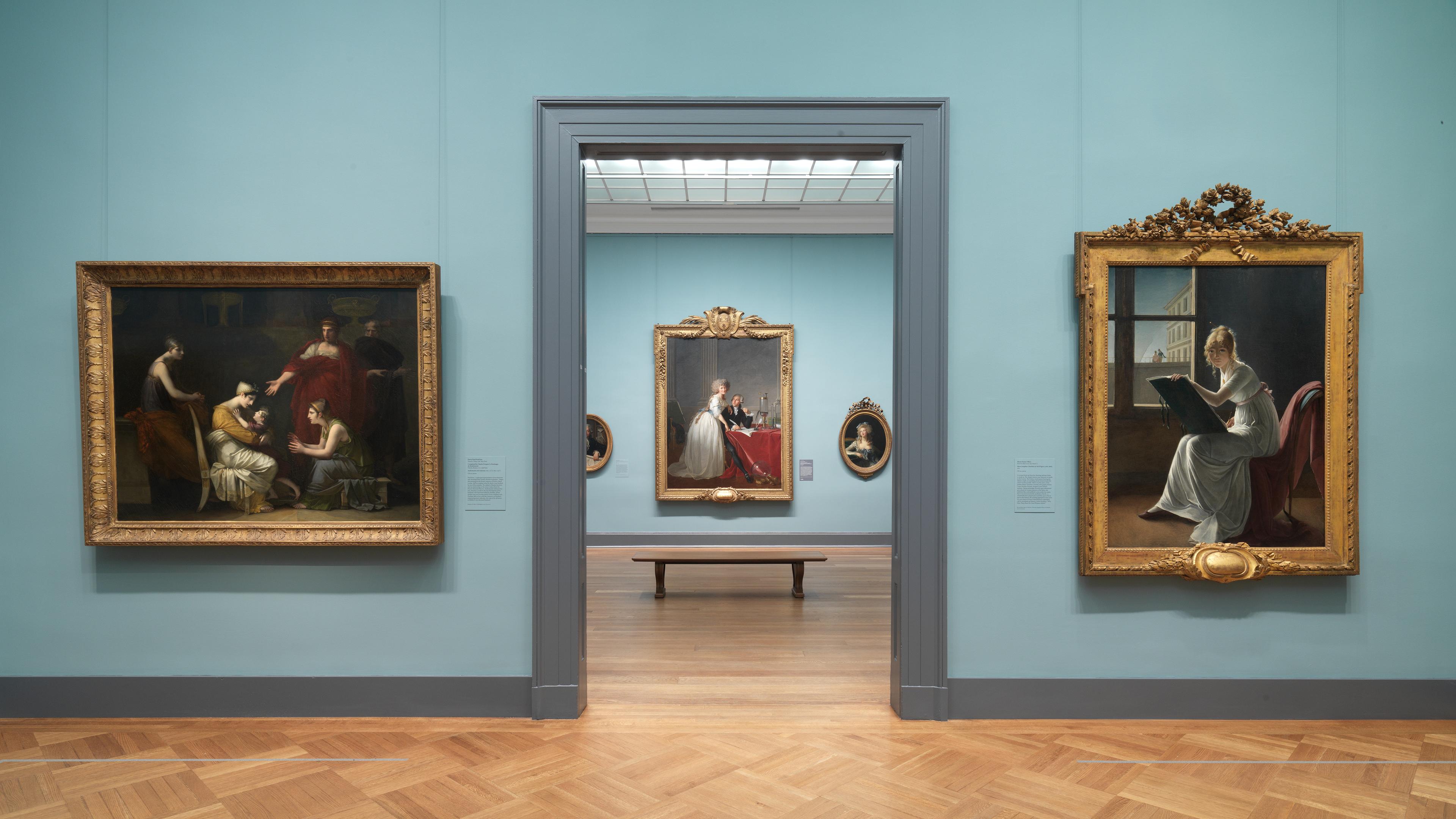 Art gallery interior featuring oil paintings of interior scenes in ornate gold frames, with a blue wall and wooden flooring.