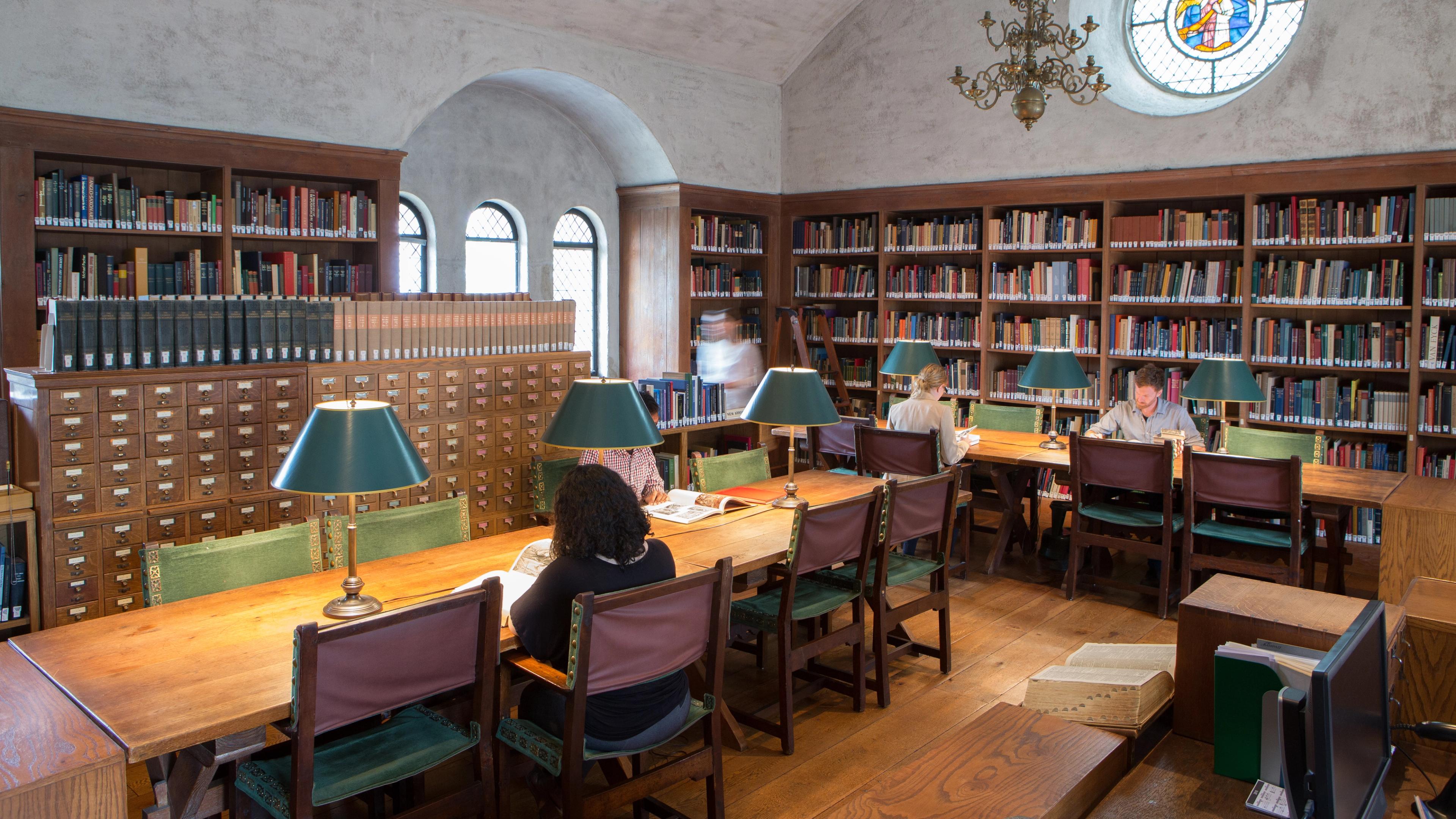 A cozy library with wooden shelves with people studying at long wooden tables illuminated by green lamps and a stained glass window above.