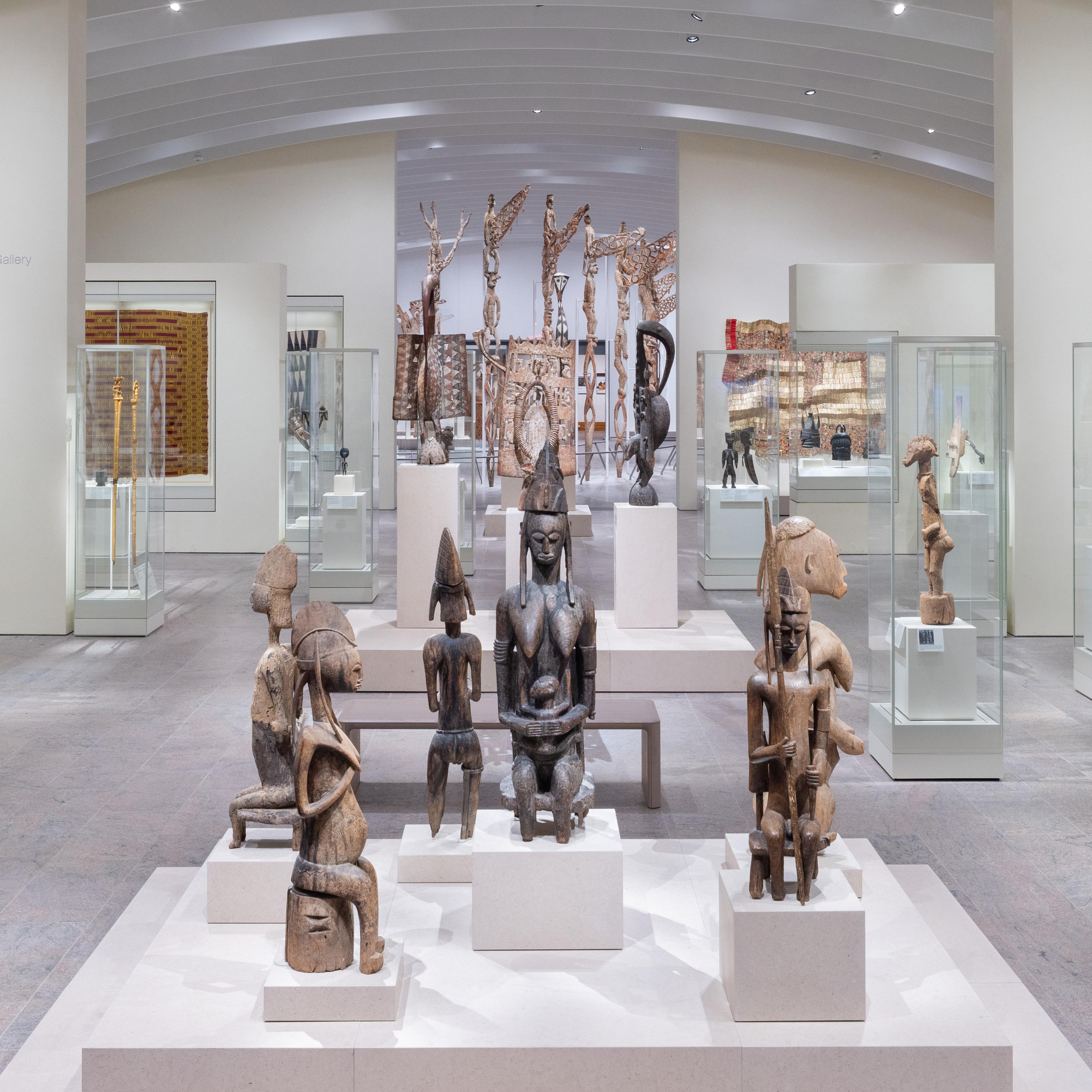 Brightly lit gallery with objects in vitrines at various elevations in the background. In the center are several seated statues on pedestals.