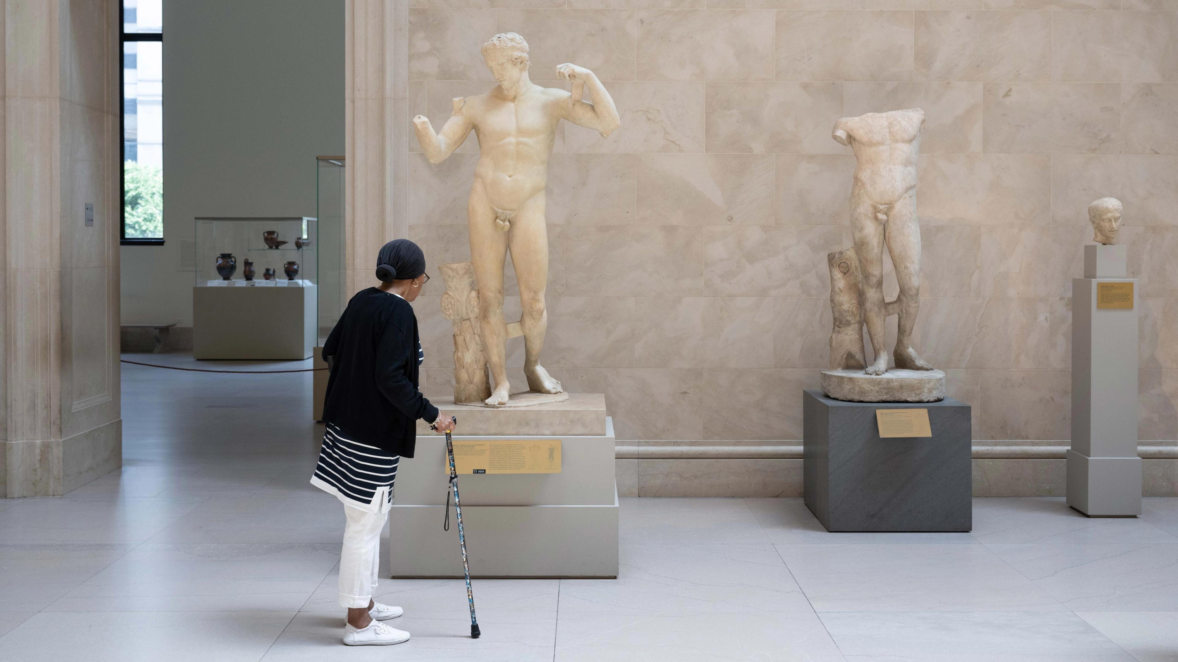 woman with a walking cane observing a Greek and Roman era marble statue.