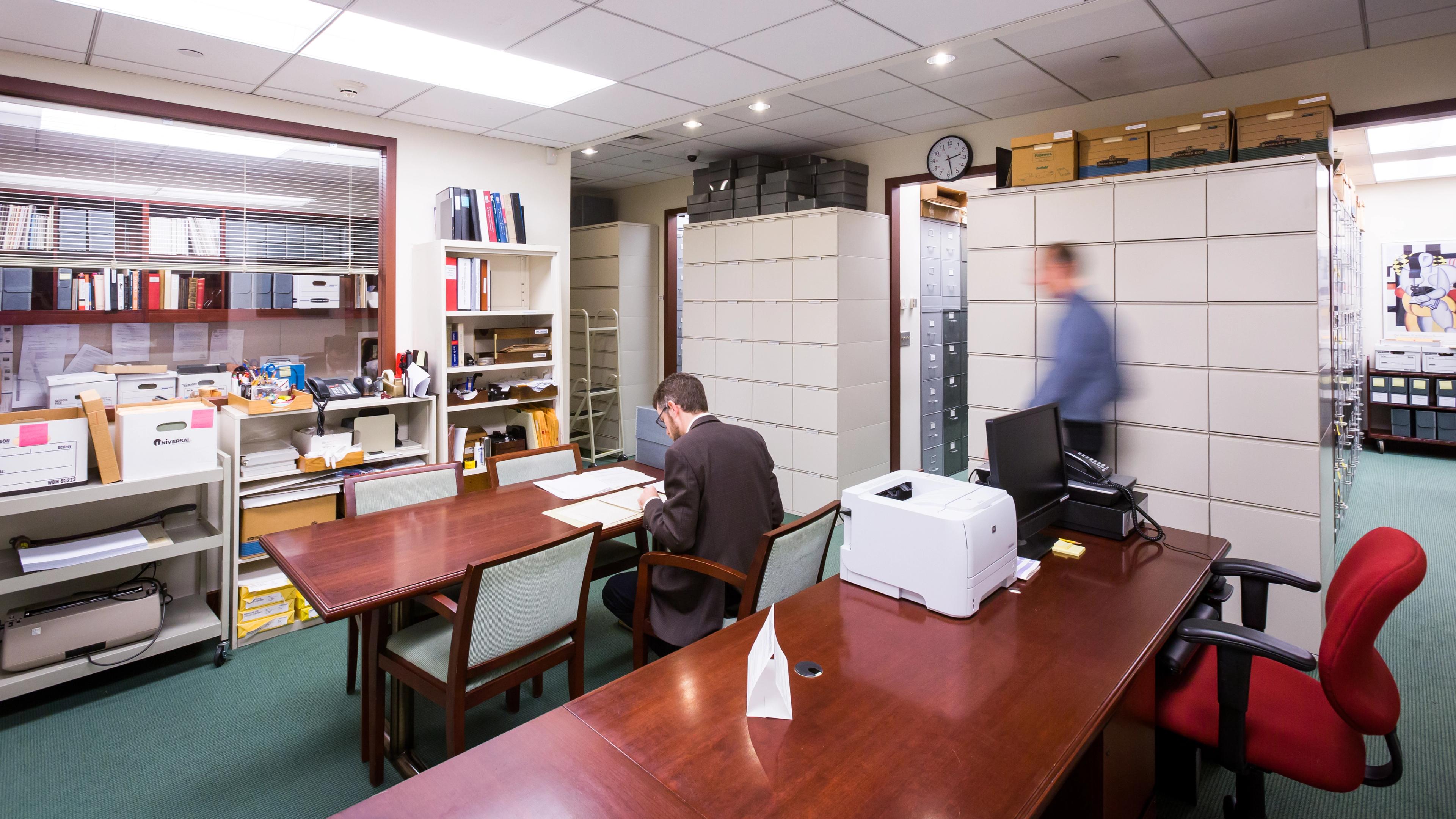 A small office space with two long wooden table, shelves filled with office supplies, and floor to ceiling filing cabinets. There is a person in a suit seated at the table looking at papers and another person walking near the filing cabinets.