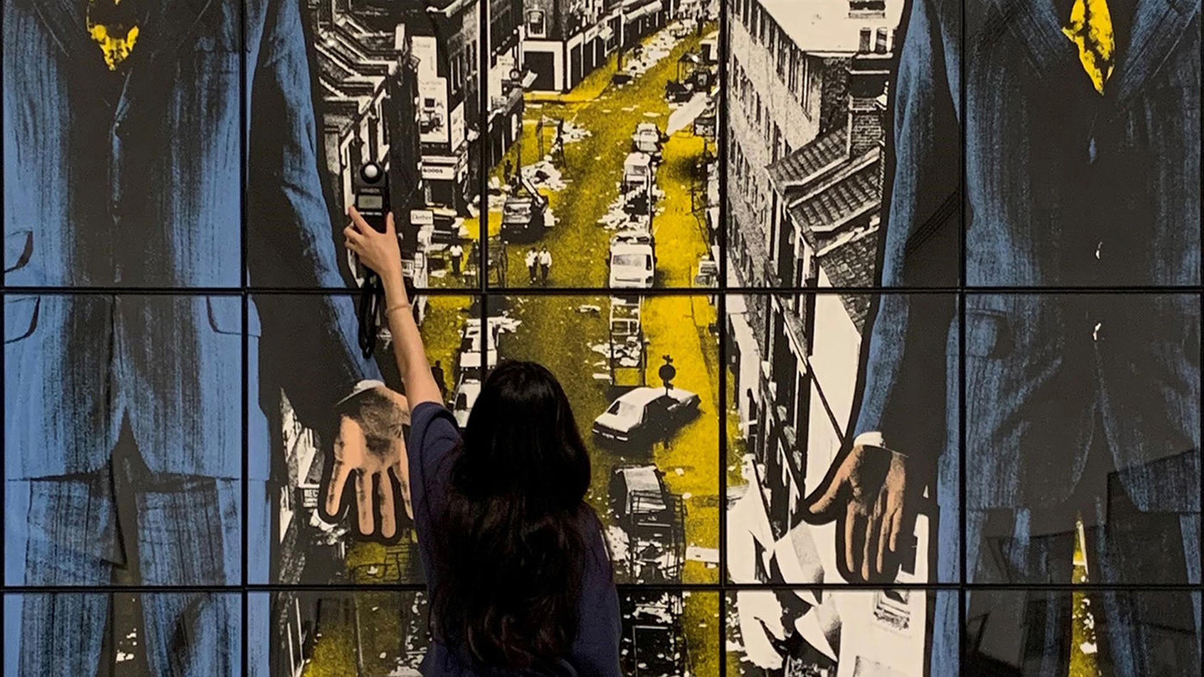 Woman facing large grid of color photographs, reaching up with small light measuring tool