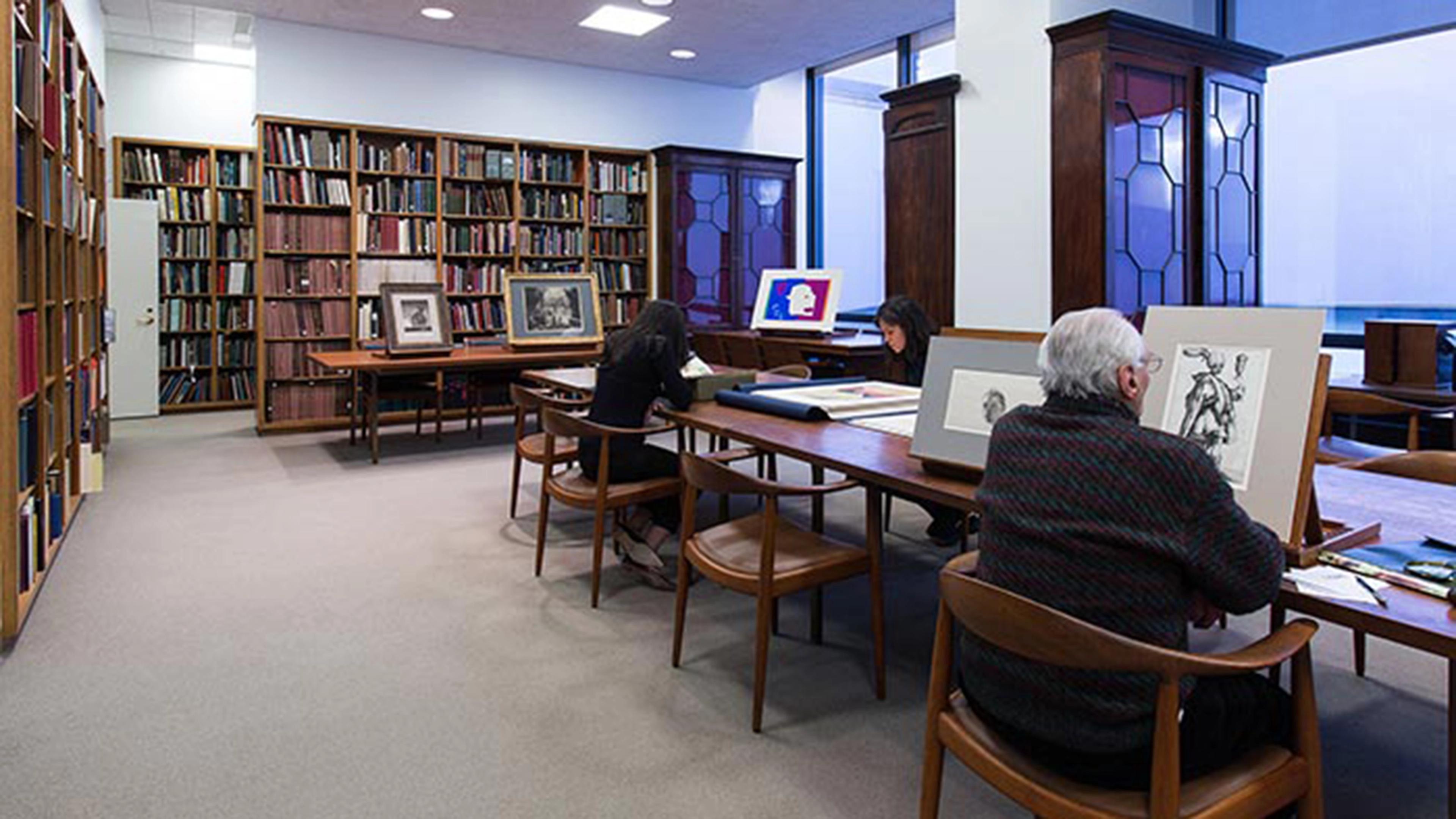 A narrow library with a long dark wood table and glass fronted cabinets with a dark-haired woman and elderly man looking at art, and in the background shelves of books