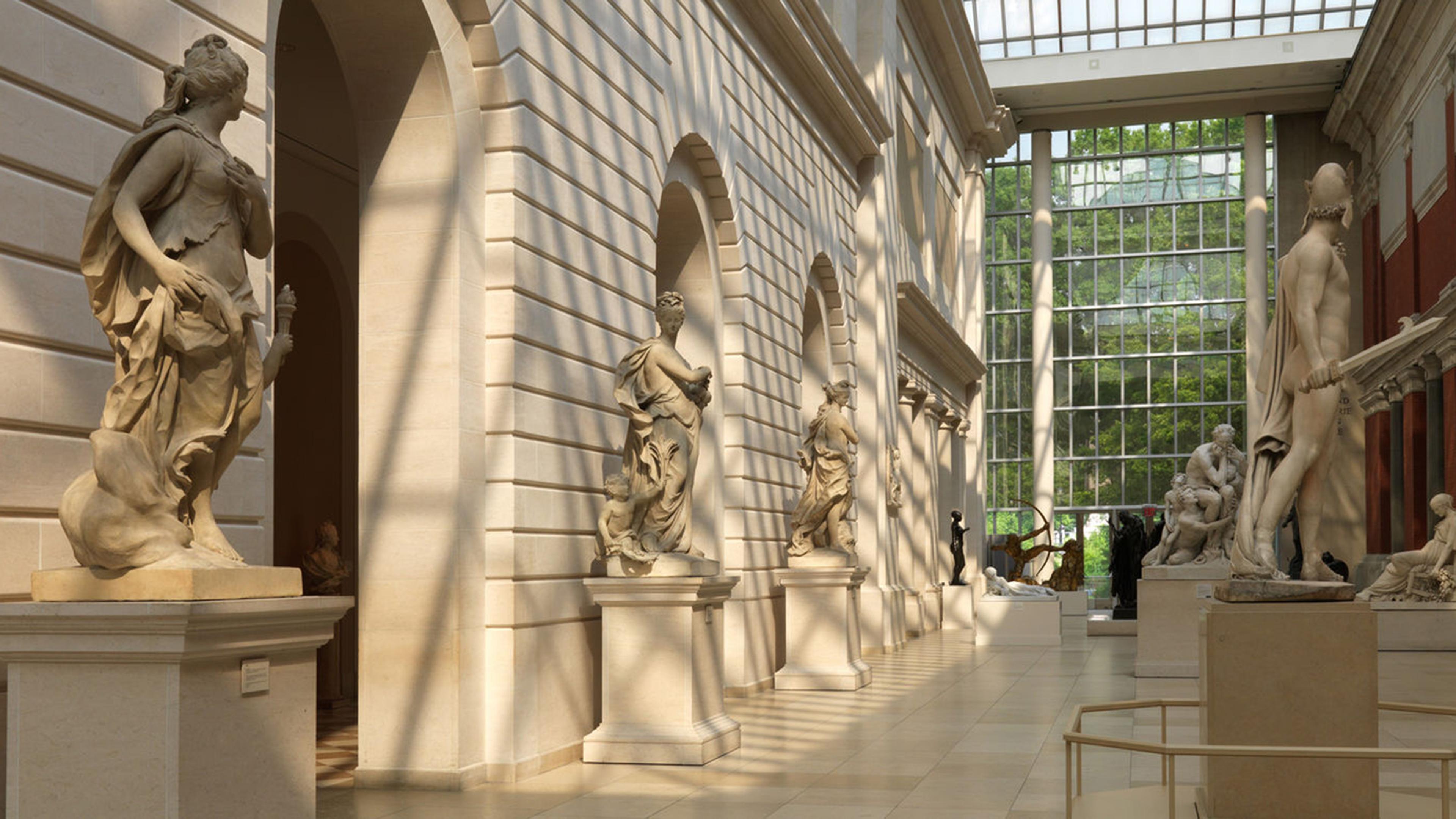 A sunlit gallery featuring classical sculptures against elegant stone walls, with large windows allowing natural light to flow in.