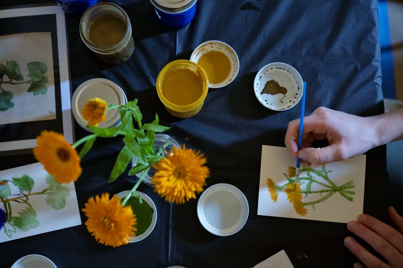 Close-up of a hand holding a paintbrush surrounded by paintings of flowers and watercolor pigments in jars.