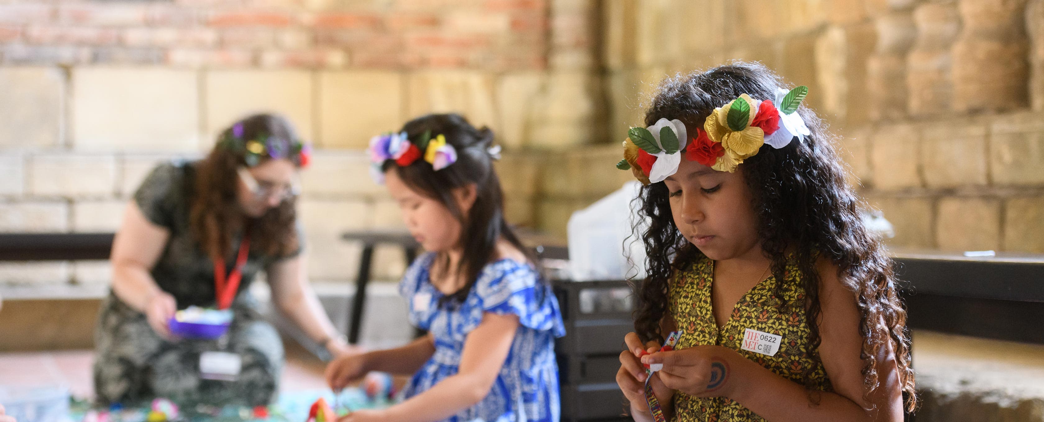 Two girls, wearing paper flower garlands in their hair engage in arts and crafts within a stone walled gallery.