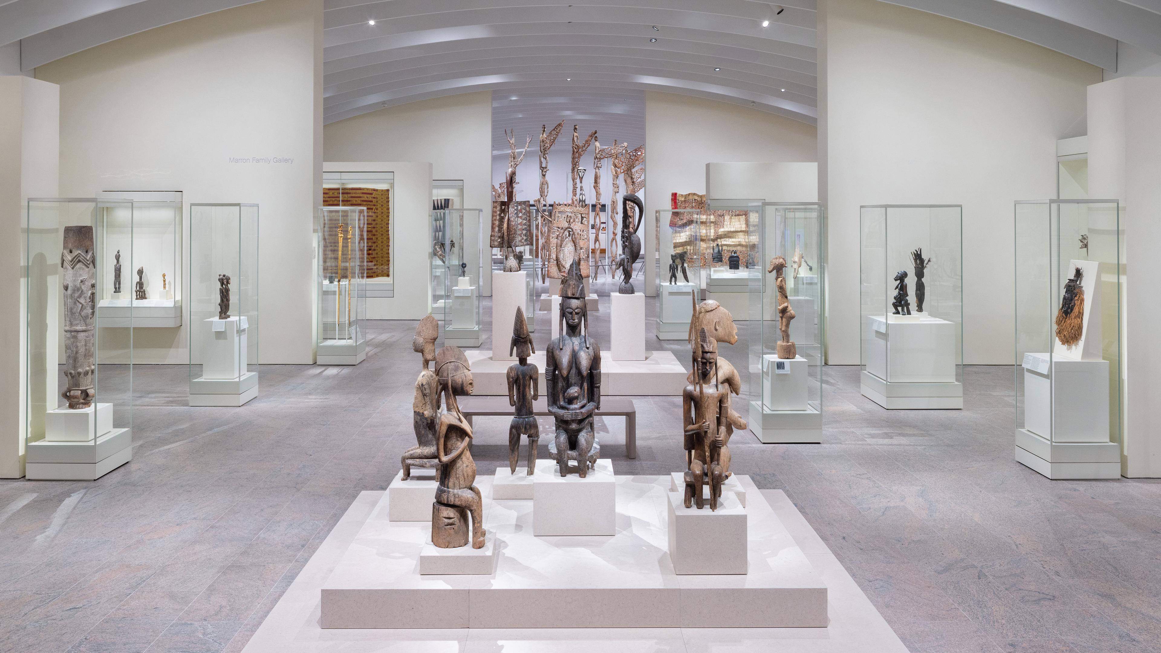 Brightly lit gallery with objects in vitrines at various elevations in the background. In the center are several seated statues on pedestals.