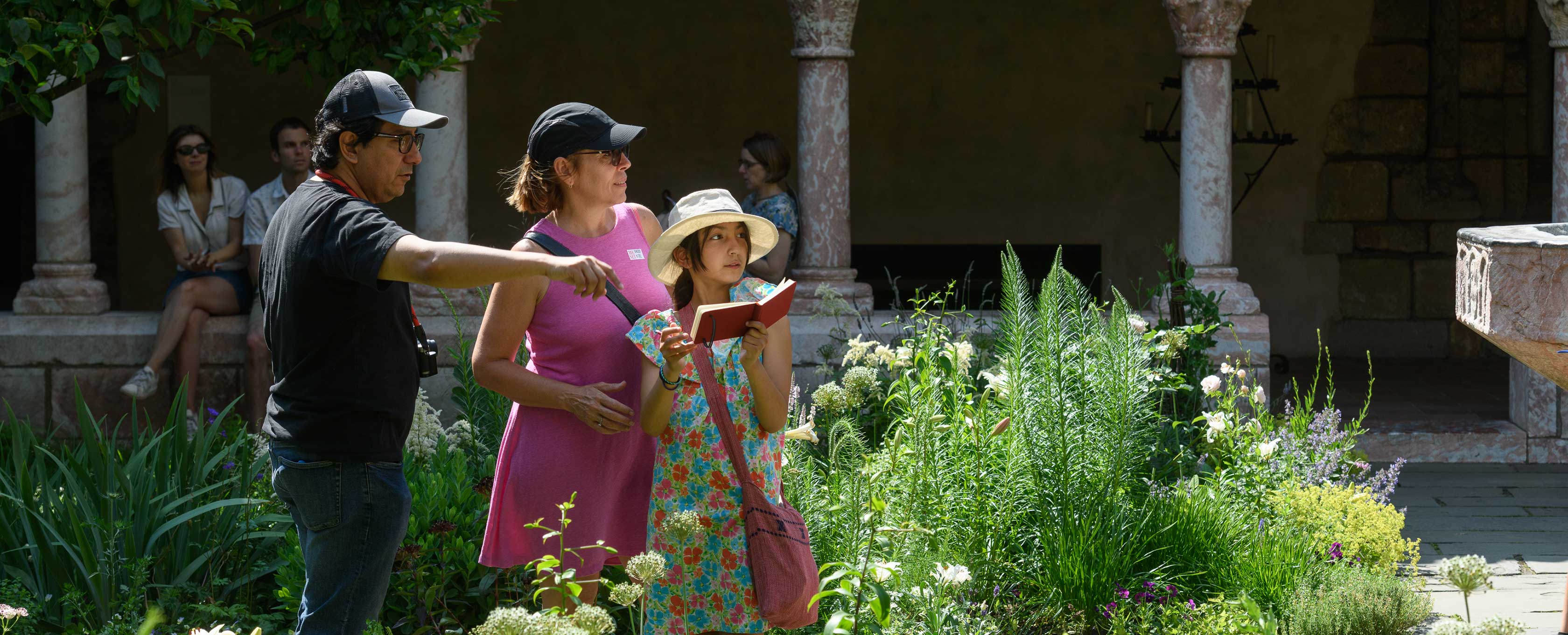 A man, woman, and a female child stand among tall grasses and plants. The child is holding a book with a red cover. They are looking at something that is out of our purview. Behind them, Met visitors are sitting between decorative marble arches.